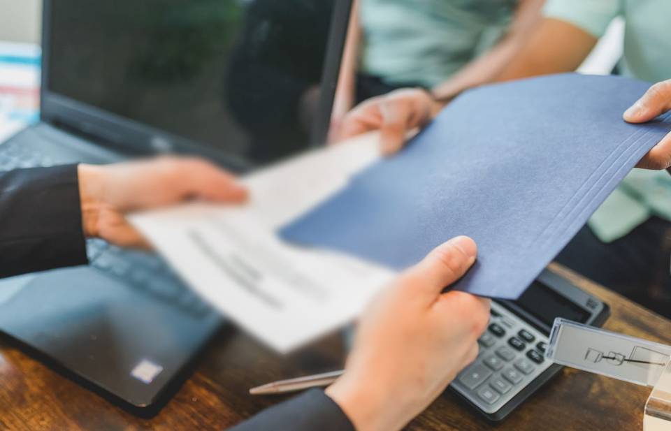 يدان تتبادلان ملفاً أزرق وأوراقاً فوق مكتب عليه حاسوب وآلة حاسبة